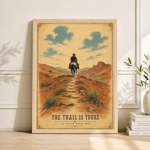 May include: A framed vintage-style print featuring a cowboy on horseback riding along a desert trail. The artwork has a sepia tone with blue sky and clouds. The text at the bottom reads "THE TRAIL IS YOURS" and "GO YOUR OWN WAY".