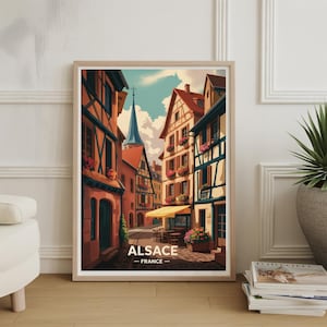 May include: A framed print featuring a colorful illustration of a European street scene. The artwork depicts buildings with red tile roofs, flower boxes, and a church steeple. The text "ALSACE - FRANCE" is displayed at the bottom.