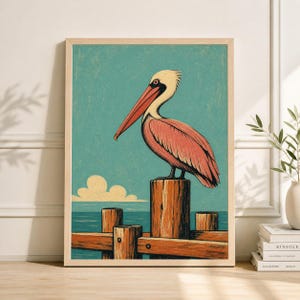Könnte beinhalten: Gerahmter Kunstdruck mit einem Pelikan auf einem Holzpfahl. Der Pelikan hat rosa und weiße Federn und einen langen roten Schnabel. Der Hintergrund zeigt einen hellblauen Himmel, Wolken und das Meer. Der Rahmen ist hellbraun.