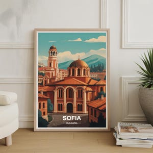 May include: Framed art print featuring a stylized illustration of Sofia, Bulgaria. The artwork depicts a church with a domed roof and a tall bell tower, rendered in warm tones of orange and brown against a blue sky. The word "SOFIA" is printed at the bottom.