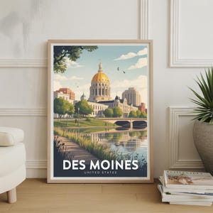 May include: Framed art print featuring a stylized illustration of Des Moines, Iowa. The artwork depicts a gold-domed building, a bridge, and a river, with the city name and "United States" at the bottom. The print is in a light wood frame.