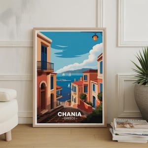 May include: Framed art print of Chania, Greece. The artwork depicts a colorful street scene with buildings in shades of orange and red, leading to a blue sea with sailboats. The sky is a bright blue with fluffy white clouds. The print is in a light wood frame.