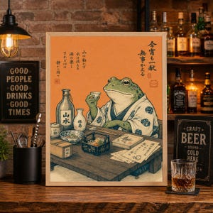 May include: A framed print featuring a frog in traditional Japanese attire, holding a cup of sake. The artwork includes Japanese text and various sake bottles and dishes on a wooden table. The background is a warm orange hue.