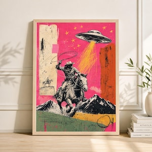 May include: A framed art print featuring a cowboy on horseback roping, with a UFO above emitting a beam of light. The background is a vibrant pink with orange and green accents, and a mountain range. The artwork has a vintage, collage style.