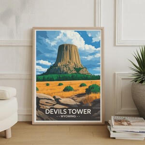 Puede incluir: Una impresión artística enmarcada con Devils Tower en Wyoming. La obra de arte representa la formación geológica en un paisaje estilizado y colorido con cielo azul y nubes. El texto "DEVILS TOWER - WYOMING -" está en la parte inferior.