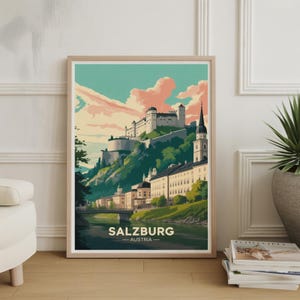 May include: A framed art print featuring a stylized illustration of Salzburg, Austria. The artwork depicts a castle atop a green hill, with buildings and a river below. The sky is a mix of blue and pink, with the word "SALZBURG" at the bottom.