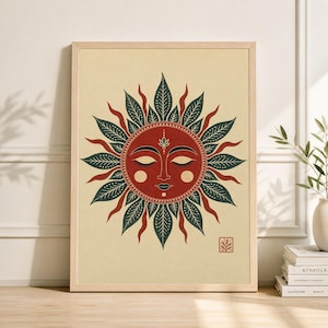 May include: A framed art print featuring a stylized sun design. The sun has a red face with closed eyes, surrounded by green and red leaf-like rays. The artwork is set against a cream-colored background, with a small square emblem in the lower right corner.