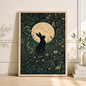 Pode incluir: Uma impressão artística emoldurada com uma silhueta preta de um coelho olhando para uma lua cheia. A obra de arte é ambientada em um campo verde escuro com elementos florais e de folhagem detalhados. A lua é de uma cor dourada suave, e a moldura é de madeira clara.