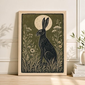May include: A framed art print featuring a black hare sitting in tall grass, with a full moon in the background. The artwork has a vintage, woodcut style, with a muted green and beige color palette. The print is in a light-colored wooden frame.