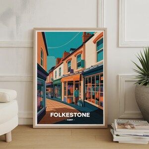 May include: A framed art print of Folkestone, Kent, showing a street scene in a stylized illustration. The artwork uses orange, teal, and white hues to depict shops and buildings. The word "FOLKESTONE" is printed in white at the bottom.