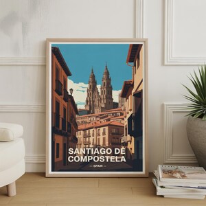 May include: Framed art print featuring a stylized illustration of Santiago de Compostela, Spain. The artwork depicts a street scene with buildings and a cathedral under a blue sky. The text "SANTIAGO DE COMPOSTELA - SPAIN" is displayed at the bottom.