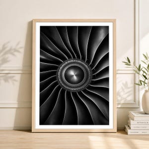 May include: A framed black and white photograph of a jet engine turbine. The image showcases the intricate design of the blades, radiating from a central hub. The artwork is displayed in a light-colored wooden frame, adding a touch of warmth to the industrial aesthetic.