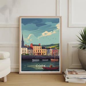 Puede incluir: Una impresión artística enmarcada con una colorida ilustración de Cork, Irlanda. La obra de arte representa una fila de edificios con fachadas coloridas a lo largo de un río, con barcos y un campanario. La impresión incluye el texto "CORK - IRELAND".