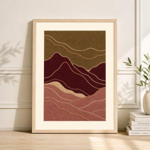 May include: Framed abstract art print featuring a textured landscape design. The artwork showcases layered mountain-like forms in shades of burgundy, brown, and pink, with gold-colored accent lines. The print is set within a light-colored wooden frame.