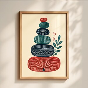 May include: A framed art print featuring a stack of patterned, oval-shaped stones in red, teal, and navy blue. The design includes a sun, leaf, and geometric shapes on a cream background. The wooden frame adds a natural touch.