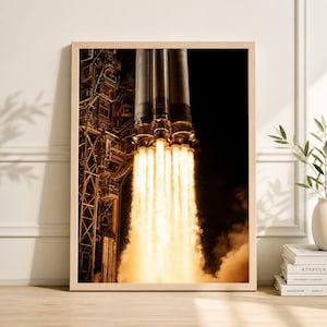 May include: A framed print of a rocket launching into space. The image shows the rocket's engines emitting bright orange flames and smoke against a black background. The rocket is mounted on a launch structure.