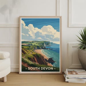 May include: A framed print of a coastal landscape with a beach, cliffs, and ocean under a blue sky with puffy white clouds. The print is labeled "- SOUTH DEVON -" in white text.