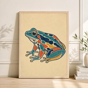 May include: A framed art print featuring a colorful illustration of a frog. The frog is depicted in shades of blue, orange, and yellow against a beige background. The artwork has a vintage, retro style, and is suitable for home decor.