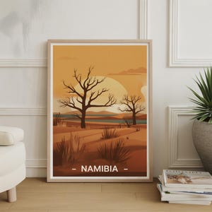 May include: Framed art print featuring a desert landscape with leafless trees under a warm, orange sky. The artwork includes a river and the word "NAMIBIA" at the bottom. The print is in a light wood frame.