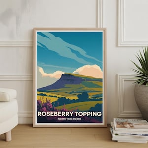 Op de afbeelding: Een ingelijste kunstdruk met een gestileerd landschap van Roseberry Topping in de North York Moors. Het kunstwerk toont een berg onder een blauwe lucht met wolken en velden in groen, geel en paars. De print bevat de tekst "ROSEBERRY TOPPING - NORTH YORK MOORS".