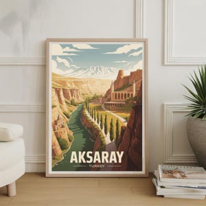 May include: A framed travel poster featuring Aksaray, Turkey. The artwork depicts a river winding through a canyon, with buildings and trees lining the banks. The poster has a light wood frame and the text "AKSARAY TURKEY" at the bottom.