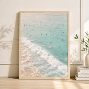 Puede incluir: Impresión enmarcada que presenta una serena escena de playa. La imagen captura un océano turquesa con numerosos surfistas, una ola blanca espumosa y una orilla arenosa. El marco de madera complementa los tonos naturales de la obra, creando una estética relajante.