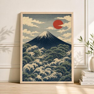 May include: A framed print of a Japanese landscape featuring Mount Fuji. The artwork depicts a snow-capped mountain, a red sun, and stylized waves and trees in shades of blue, green, and white. The print is in a light-colored wooden frame.
