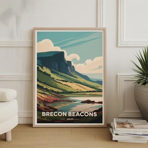 May include: A framed art print featuring a landscape scene of the Brecon Beacons in Wales. The artwork showcases a mountain range, a lake, and a sky with clouds. The print is in a wooden frame and is displayed on a wooden floor.
