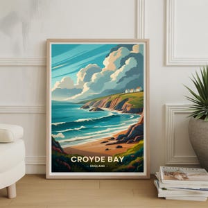 May include: A framed art print of Croyde Bay, England. The artwork depicts a coastal scene with a sandy beach, turquoise waves, and a green cliffside under a blue sky with fluffy white clouds. The print is in a light wood frame.