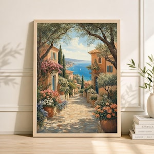May include: A framed print of a European coastal town. The artwork depicts a cobblestone street lined with buildings, flowers, and trees. The buildings have orange-tiled roofs and blue shutters. The background shows the sea and sky.