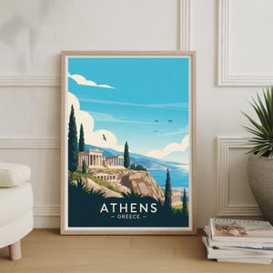 May include: A framed art print featuring a stylized illustration of Athens, Greece. The artwork depicts a classical building on a cliff overlooking the sea, under a blue sky with clouds. The print is in a light wood frame and includes the text "ATHENS - GREECE".