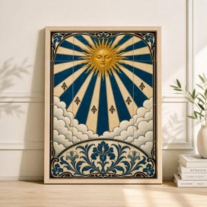 Op de afbeelding: Een ingelijste kunstdruk met een gestileerde zon met een gezicht, die licht uitstraalt tegen een blauwe en crèmekleurige achtergrond. Het kunstwerk bevat decoratieve wolken, fleur-de-lis en bloemelementen in blauwe en crèmetinten, in een licht houten frame.