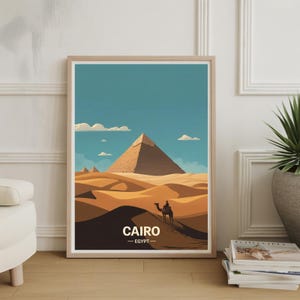 Może przedstawiać: Oprawiony wydruk artystyczny przedstawiający stylizowaną ilustrację piramid w Kairze, w Egipcie. Grafika przedstawia jeźdźca na wielbłądzie przemierzającego pustynny krajobraz pod błękitnym niebem z chmurami. Słowo "CAIRO" jest wydrukowane na dole.
