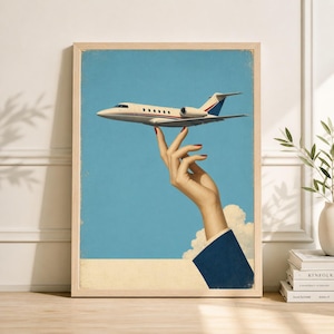 May include: A framed print featuring a vintage-style illustration of a hand holding a small airplane against a blue sky. The airplane is white with red and blue accents. The hand has red nail polish.