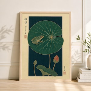 Pode incluir: Impressão artística emoldurada com um sapo verde em uma grande folha de nenúfar. A obra de arte tem um fundo azul escuro com uma borda bege clara. Uma flor de lótus vermelha e texto japonês também são visíveis.