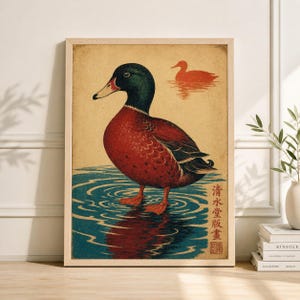 May include: A framed art print depicting a mallard duck in water, with a red body, green head, and yellow beak. A second duck is in the background. The artwork is in a vintage style with Japanese text. The print is in a wooden frame.