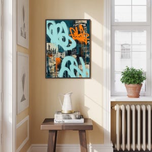 May include: Framed abstract art print featuring graffiti-style designs in light blue and orange against a dark teal background. The artwork is displayed in a room with a wooden side table, a white pitcher, and a window with a potted plant.