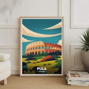 May include: A framed art print featuring a stylized illustration of the Pula Arena in Croatia. The artwork depicts the ancient Roman amphitheater in shades of beige, orange, and red against a blue sky with white clouds. The word "PULA" is printed below the image.