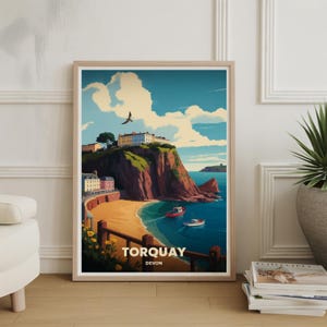 May include: A framed art print featuring a coastal landscape of Torquay, Devon. The artwork depicts a beach, boats, and buildings atop a cliff, under a blue sky with clouds. The print includes the text "TORQUAY DEVON".