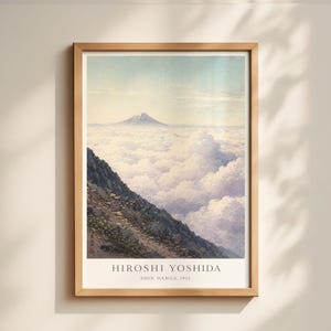 May include: Framed print of a mountain landscape with a snow-capped peak rising above a sea of clouds. The artwork, by Hiroshi Yoshida, features a rocky hillside in the foreground. The print is in a light wood frame.