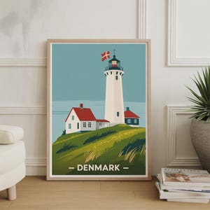 May include: Framed art print featuring a white lighthouse with a red-roofed building and a Danish flag. The artwork has a minimalist style with a blue sky and green grassy hill. The word "DENMARK" is printed at the bottom.