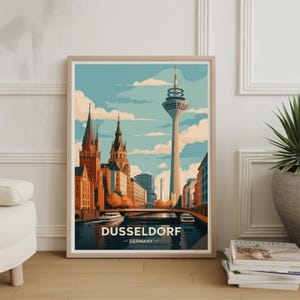 May include: A framed art print featuring a stylized illustration of Düsseldorf, Germany. The artwork depicts the city's skyline with the Rheinturm tower, buildings, and a river with boats. The print has a light wood frame and the text "DUSSELDORF - GERMANY".