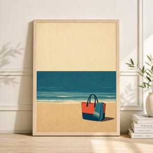 May include: A framed art print depicts a beach scene. The artwork features a tan sky, a blue ocean, and a sandy beach. A red and blue tote bag and a blue towel are on the beach. The print is in a light wood frame.