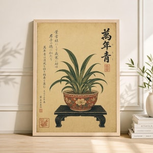 Puede incluir: Una impresión botánica enmarcada que presenta una planta en maceta con largas hojas verdes y una maceta con estampado floral rojo y blanco. La impresión tiene texto japonés y está sobre un fondo beige. El marco es de madera clara.