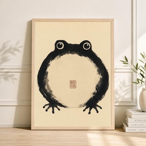 Puede incluir: Una impresión artística enmarcada con un diseño minimalista de rana negra. La rana tiene un cuerpo circular y ojos grandes, con un pequeño sello rojo en el centro. La obra de arte está sobre un fondo color crema.