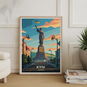 May include: Framed art print featuring a stylized illustration of a monument in Kyiv, Ukraine. The artwork depicts a statue holding a Ukrainian flag against a sunset sky. The print includes the city name "KYIV" at the bottom.