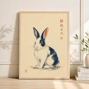 Op de afbeelding: Een ingelijste kunstdruk met een aquarelillustratie van een konijn. Het konijn heeft witte vacht met donkerblauwe vlekken en roze gevoerde oren. De achtergrond is lichtbeige, met Japanse tekst.