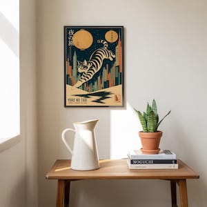 Könnte beinhalten: Gerahmter Kunstdruck mit einer stilisierten Katze, die über eine Stadtlandschaft springt. Das Kunstwerk hat eine Vintage-Ästhetik mit einem dunkelblauen Hintergrund, einer gelben Laterne und dem Text "Yoru No Tabi". Die Katze ist schwarz-weiß gestreift.