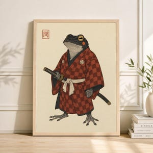 May include: A framed art print featuring a frog samurai in a red checkered kimono, holding a katana. The frog has a serious expression and is set against a cream-colored background. The print includes Japanese text in the upper left corner.