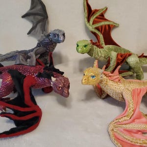 May include: Four plush dragon toys in different colors. One is dark gray with purple scales, one is red with black wings, one is light green with dark red wings, and one is gold with pink wings. Each dragon has textured scales and wings.